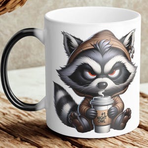 May include: White ceramic coffee mug featuring a cartoon raccoon wearing a brown hoodie and holding a coffee cup. The raccoon has a stern expression and the coffee cup reads "#1 NIGHT OWL". The mug has a black handle.