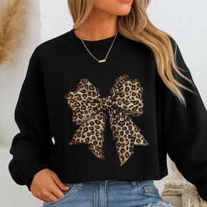 Peut inclure: Un sweat-shirt noir à manches longues arborant un grand motif central représentant un nœud avec un imprimé léopard marron et noir. Le sweat-shirt présente une encolure ronde classique et une coupe décontractée avec un motif animalier.