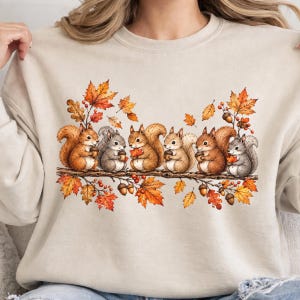 May include: A beige crewneck sweatshirt featuring a graphic illustration of six squirrels perched on a tree branch. The squirrels are depicted in shades of brown and grey, holding acorns and autumn leaves, surrounded by orange and yellow fall foliage.