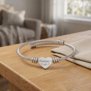 May include: A silver-toned cable wire bangle bracelet featuring a central heart-shaped charm. The charm is engraved with the text Mommy's Angel and includes a small wing graphic. The bracelet has a bypass design with metallic coil accents.