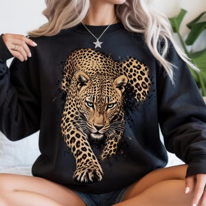 May include: A black long-sleeve sweatshirt featuring a large, detailed graphic print of a leopard in the center. The leopard is depicted in natural tan and black spotted colors with piercing blue eyes, surrounded by black paint splatter effects.