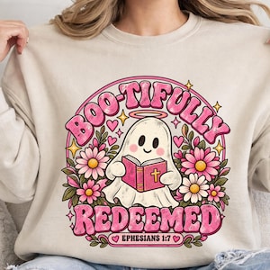 May include: A beige crewneck sweatshirt featuring a graphic of a white ghost with a halo reading a pink book. The text reads BOO-TIFULLY REDEEMED EPHESIANS 1:7. The design includes pink flowers, green leaves, small yellow stars, and pink hearts.