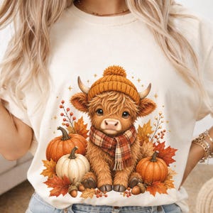 Può includere: Una t-shirt color crema con la grafica di una mucca Highland pelosa che indossa un berretto in maglia giallo senape e una sciarpa a quadri. La mucca è circondata da zucche arancioni e bianche, foglie autunnali, bacche rosse e ghiande.