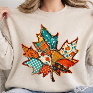 May include: A beige crewneck sweatshirt featuring a large central graphic of a maple leaf composed of various patchwork patterns. The leaf design includes sections of plaid, gingham, floral, and pumpkin prints in shades of orange, teal, and white.