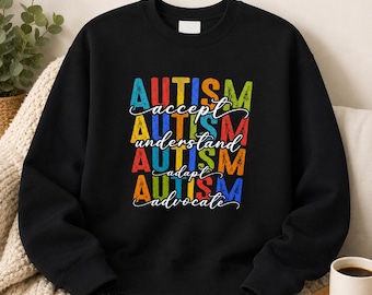 May include: A black crewneck sweatshirt featuring a multi-colored graphic print. The design displays the word AUTISM in distressed block letters, alternating with the cursive words accept, understand, adapt, and advocate in white script.