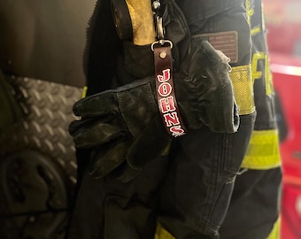 May include: A black leather keychain with the name "JOHNS" printed in red letters. The keychain is attached to a silver ring and a black and yellow fire fighter jacket.