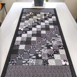 May include: A black and white patchwork table runner with a checkerboard design. The runner features squares of various patterns, including polka dots, floral, and geometric shapes. A solid black border frames the runner, and a dotted pattern adorns the bottom edge.