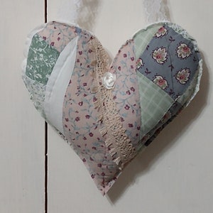 May include: A patchwork heart-shaped pillow with a floral pattern in shades of pink, green, and blue. The pillow has a white lace trim and a white button in the center. It is hanging from a white lace ribbon.