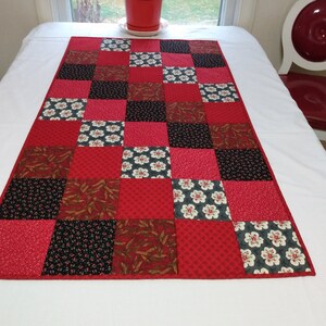 May include: Red quilted table runner with a patchwork design featuring various red, black, and white floral patterns. The runner is made of fabric squares sewn together.