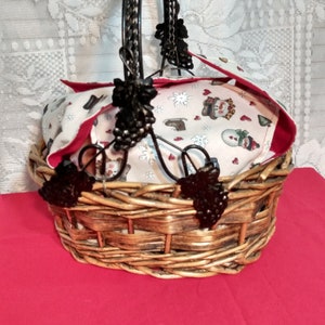 May include: A wicker basket with a black metal handle and decorative accents. The basket is lined with a white and pink fabric with a winter-themed print featuring snowmen, hearts, and snowflakes.