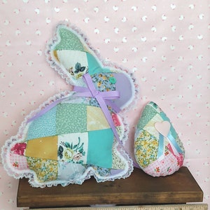 May include: A patchwork Easter bunny and egg, crafted from fabric squares in pastel colors and floral patterns. The bunny has a lavender bow and lace trim. The egg features a pink heart and ribbon. The objects are displayed on a small wooden shelf.