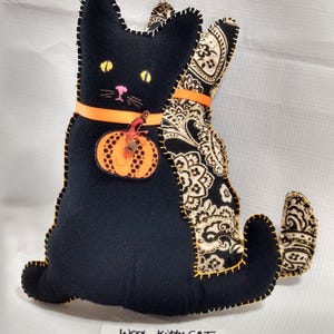 May include: A black wool cat-shaped plush toy with yellow eyes, a pink nose, and orange ribbon. The cat has a pumpkin applique and a paisley patterned back. The edges are stitched with yellow thread. The text on the image reads "Wool Kitty Cat Tail Right, BK+Tan Paisley Back."