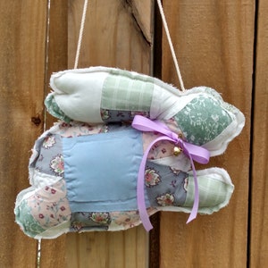May include: A handmade fabric bunny decoration with a patchwork design in pastel colors. The bunny has a light blue square on its back and a lavender ribbon with a small bell. It hangs from a white string against a wooden fence.