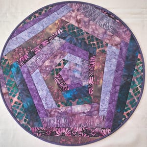 May include: A circular fabric mat featuring a geometric pentagonal pattern constructed from various quilted strips. The design incorporates shades of purple, lavender, and teal with batik-style floral and grid motifs, finished with a purple border.