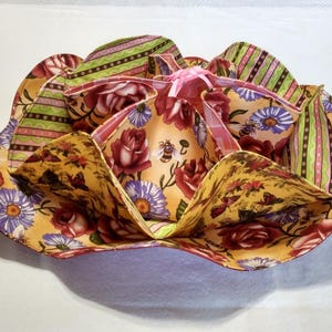 May include: A multi-layered fabric bowl with a floral design. The bowl features a variety of patterns, including roses, bees, butterflies, and stripes. The colors include yellow, red, purple, green, and pink. The bowl is designed to resemble a flower.