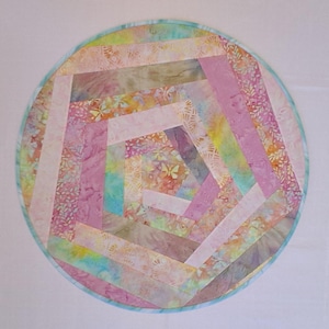 May include: A circular fabric mat featuring a spiral patchwork design composed of strips in shades of pink, lavender, yellow, and teal. The fabric displays various patterns including small butterflies, leaves, and mottled textures, finished with a thin teal border.