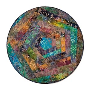 May include: A circular fabric mat featuring a spiral pattern of batik-style strips in shades of blue, green, purple, orange, and yellow. The textile surface displays various floral and abstract patterns across the multicolored, layered fabric segments.