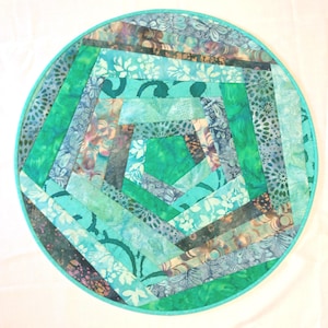 May include: A round, handmade quilt with a teal and green color scheme. The quilt features a geometric design with various fabric patterns, including floral and abstract prints. The outer edge is a solid teal, and the diameter is approximately 12 inches.