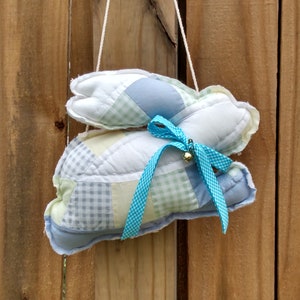 May include: Two stuffed bunny-shaped ornaments hanging from a white string. The bunnies are made of patchwork fabric in pastel colors, including blue, green, and yellow. A blue ribbon with a small bell is tied around the center of the bottom bunny. The background is a wooden fence.