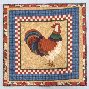 May include: A quilted placemat with a red, white, and blue checkered border and a rooster design in the center. The rooster is brown, white, and purple with a green and brown comb and wattle.
