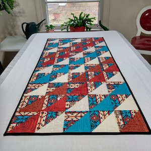 May include: A table runner with a geometric pattern of red, white, and blue triangles. The runner is made of fabric and has a black border.