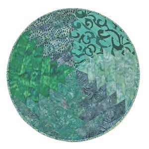 May include: A circular fabric mat featuring a geometric pattern of pieced fabric strips in various shades of green, teal, and blue. The design incorporates batik-style prints with floral and abstract motifs, finished with a thin patterned border.