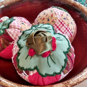 May include: Three fabric pincushions in a brown bowl. The pincushions are covered in patchwork fabric with floral and plaid patterns. One pincushion has a green leaf design with a brown satin bow and red felt flowers.
