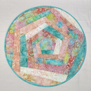 May include: A round, colorful quilted placemat with a geometric design. The placemat features a central pentagon shape surrounded by radiating strips of fabric in shades of pink, teal, peach, and white. A teal border frames the design.