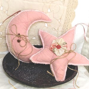 May include: Two pink fabric ornaments, a crescent moon and a five-pointed star, with a rustic twine bow. The star has a floral embellishment with a red button in the center.