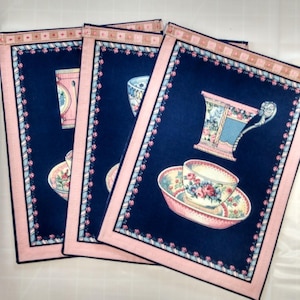 May include: Four navy blue placemats with a pink border and floral teacup designs. Each placemat features a different teacup and saucer illustration in shades of pink, blue, and yellow. The border has a repeating floral pattern.