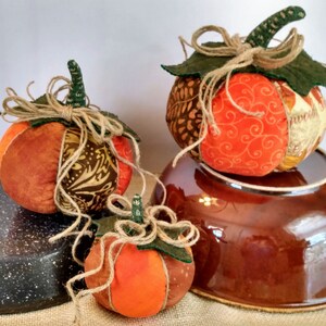 May include: Three handmade fabric pumpkins in orange, brown, and green with burlap ribbon and leaves. The pumpkins are sitting on a brown bowl and a black speckled surface.