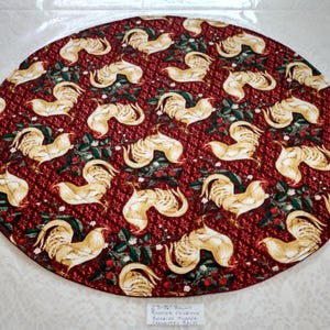 May include: A round, 23.75-inch rooster-themed table topper. The topper features a repeating pattern of cream-colored roosters on a deep red background with green leaves and red berries. The edge is finished with a stitched border.