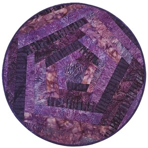 May include: A circular fabric mat featuring a spiral pentagonal pattern constructed from various shades of purple and violet batik cotton strips. The design radiates from a central pentagon, with each layer showcasing distinct textures and patterns.