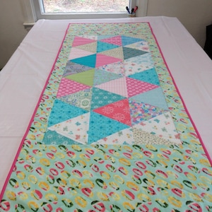 May include: A rectangular table runner with a patchwork design. The center features colorful triangles in pink, blue, and green. The border is a mint green with a pattern of watermelon rings, and a pink trim. The runner measures 19 x 46 inches.
