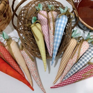 May include: A collection of fabric carrot decorations in various colors and patterns, including gingham, floral, and solid orange. Each carrot has green fabric leaves and a brown ribbon. Some carrots are in a woven basket.