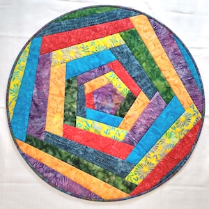 May include: A round, colorful quilted placemat with a geometric design. The placemat features a variety of vibrant colors, including red, yellow, blue, green, orange, and purple. The design is made up of rectangular fabric pieces arranged in a circular pattern.