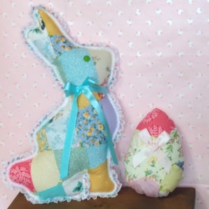 May include: A patchwork Easter bunny and egg. The bunny is made of various floral patterned fabrics in pastel colors, trimmed with white lace and a blue ribbon. The egg is similarly patterned with a pink top and a white bow.