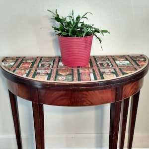 May include: A half-round wooden table with a patterned fabric top. The table has three legs and is decorated with a green and red floral pattern. A red pot with a green plant sits on top of the table.