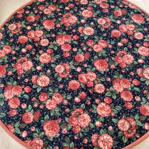 May include: A round, quilted placemat with a floral pattern. The design features large, pink roses and green leaves on a navy blue background. The edge is trimmed with a pink border. The placemat is likely made of fabric and is designed for use on a table.