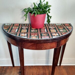 May include: A half-round wooden table with a green and brown patterned tablecloth. A red pot with a green plant sits on the table.
