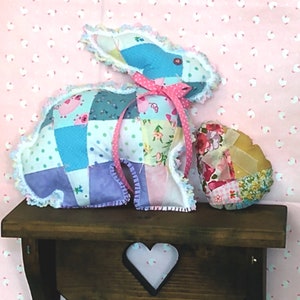 May include: A patchwork bunny and egg decoration. The bunny is made of various fabric squares in blue, purple, pink, and green, with a pink ribbon. The egg is also patchwork, with floral and solid fabrics. Both are trimmed with lace.