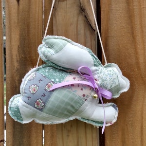 May include: A patchwork bunny decoration with a lavender ribbon and a small gold bell. The bunny is made of various floral and patterned fabrics in shades of green, white, and pink. It hangs from a white string against a wooden fence.