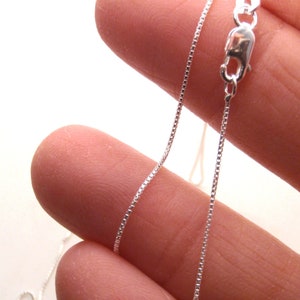 May include: A close-up of a delicate silver box chain necklace with a lobster clasp. The chain is held between fingers, showcasing its fine, square links. The clasp is shiny and reflective, set against a soft, neutral background.