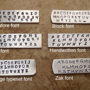 May include: Six metal tags showcasing different font styles: Pinafore, Block, Fun, Handwritten, Vintage Typeset, and Zak. Each tag displays the alphabet, with some including numbers and symbols. The tags are arranged on a brown surface.