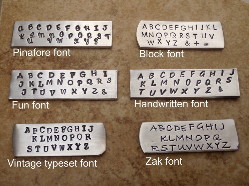 May include: Six metal tags showcasing different font styles: Pinafore, Block, Fun, Handwritten, Vintage Typeset, and Zak. Each tag displays the alphabet, with some including numbers and symbols. The tags are arranged on a brown surface.