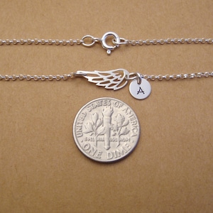 May include: A silver-colored bracelet featuring a delicate wing charm and a small round pendant with the letter "A". The bracelet is made of a fine chain and has a clasp closure. The bracelet is shown next to a dime.