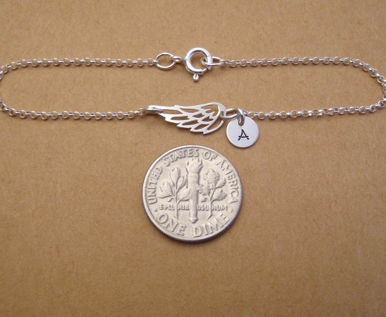 May include: A silver-colored bracelet featuring a delicate wing charm and a small round pendant with the letter "A". The bracelet is made of a fine chain and has a clasp closure. The bracelet is shown next to a dime.