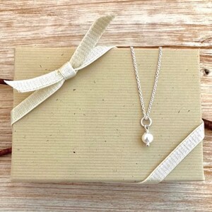 May include: A silver necklace with a small, round, white pearl pendant rests on a beige gift box tied with a cream-colored ribbon. The box has a textured surface.