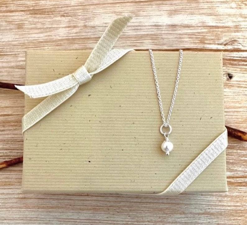 May include: A silver necklace with a small, round, white pearl pendant rests on a beige gift box tied with a cream-colored ribbon. The box has a textured surface.