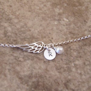 May include: A silver-toned necklace featuring a delicate wing charm, a small circular pendant with the letter "R", and a single pearl. The chain is fine and the design is simple and elegant.